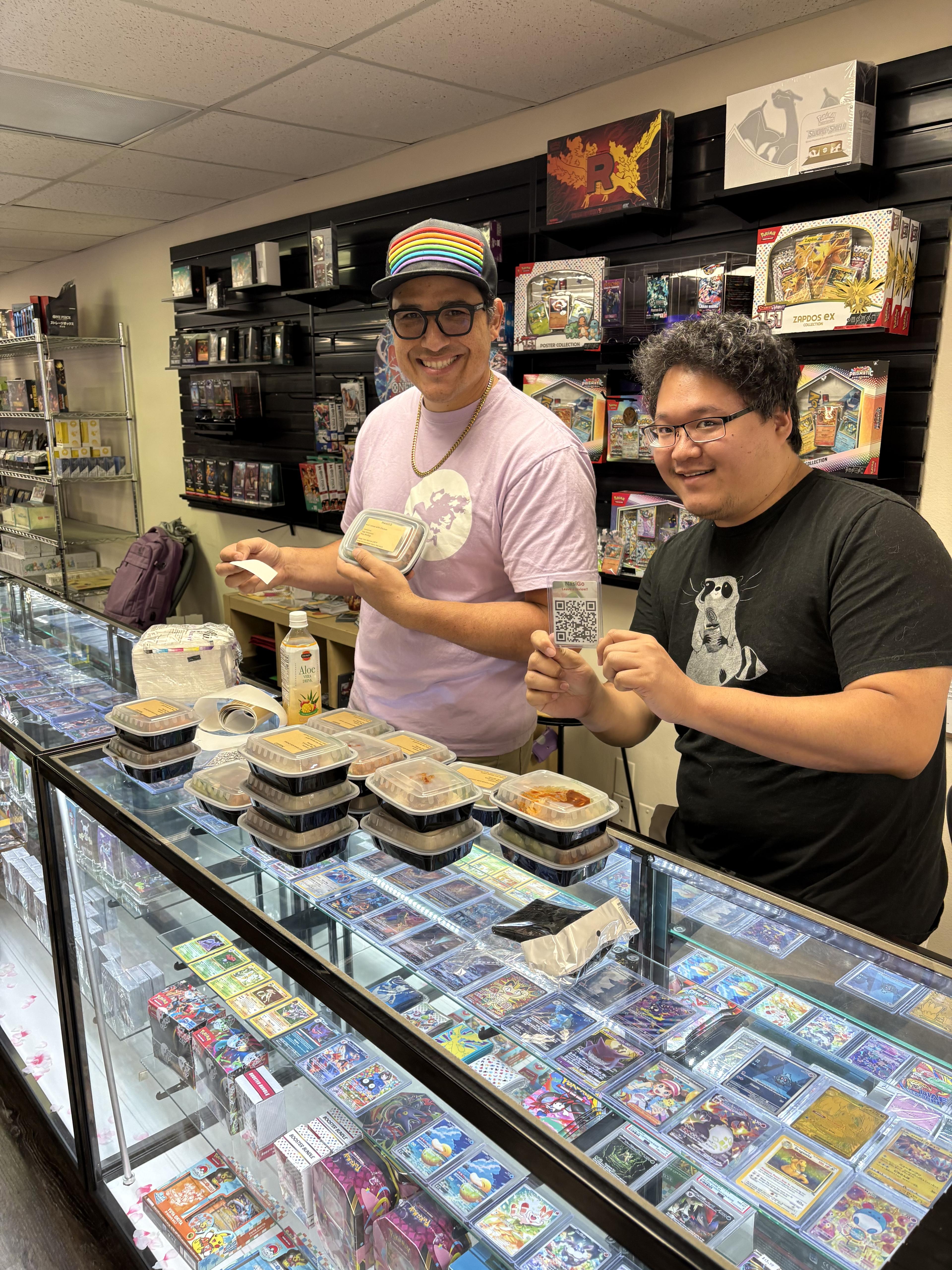 Two customers at Fei's Games holding a nasiGO box and the review QR card, with boxed meals lined up on the counter